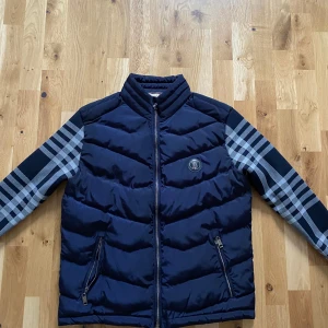 Blå pufferjacka från Burberry, S - Säljer en snygg mörkblå pufferjacka från Burberry med klassiska rutiga ärmar och foder. Jackan har dragkedja framtill, två fickor med dragkedja och ikonisk Burberry-logga på bröstet. Perfekt för dig som vill sticka ut med stilren design och lyxiga detaljer.
