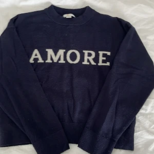 Mörkblå stickad tröja  - Mysig mörkblå stickad tröja från Gina Tricot med vit text 'AMORE' framtill. Tröjan har rund halsringning och långa ärmar, perfekt för kyliga dagar. Enkel och stilren design som passar till jeans eller kjol.