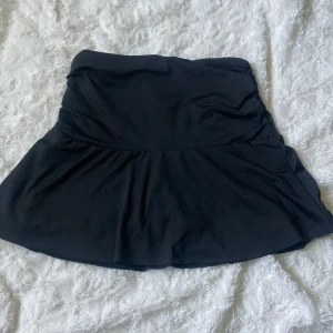 Svart sportig minikjol - Svart kortkjol i stretchigt material med inbyggda shorts under. Kjolen har en lätt utsvängd form och rynkade detaljer upptill i midjan. Perfekt för en aktiv och trendig look.