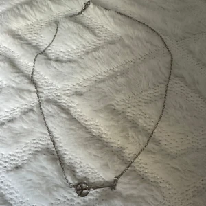 Silverfärgat halsband med nyckelberlock - Snyggt halsband i silverfärg med en tunn kedja och en berlock formad som en nyckel med peace-symbol. Perfekt accessoar för dig som gillar unika detaljer och vill sticka ut lite extra.
