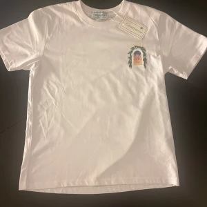 Vit t-shirt - Vit t-shirt  med färgstarkt tryck av byggnad och blad på bröstet och ryggen. Klassisk passform med rund hals och korta ärmar. Materialet är mjuk bomull och trycket har detaljer i grönt, blått, orange och gult. Perfekt för dig som gillar stilrena men unika plagg.OBS INTE ÄKTA