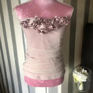 Ljusrosa bandeau tubtopp - Supersöt ljusrosa bandeau tubtopp med stora tygblommor längs överkanten. Toppens tyg är lätt transparent och har en draperad look som ger en snygg och feminin siluett. Perfekt för dig som vill sticka ut med en romantisk vibe.
