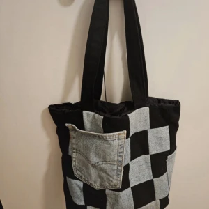 Rutigt shoppingväska i jeans och svart tyg - Unik shoppingväska med rutigt mönster i svart tyg och ljusblå jeans. Framsidan har en cool ficka av återvunnen jeans, perfekt för mobilen eller nycklar. Väskan har breda axelband och är rymlig nog för allt du behöver till skolan eller stan.