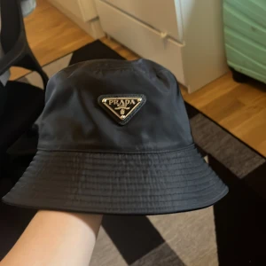Svart bucket hat från Prada - Snygg svart bucket hat från Prada med ikonisk metallogo framtill. Hatten är tillverkad i ett glansigt syntetmaterial som ger en lyxig känsla och passar perfekt till streetwear eller för att lyfta din outfit. Klassisk form med bredare brätte och diskret sömnad.