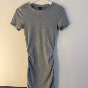 Grå ribbad kortklänning S - Säljer en grå ribbad kortklänning från Divided i storlek S. Klänningen har korta ärmar, rund halsringning och snygg rynkdetalj på sidan som ger en cool twist. Perfekt för dig som gillar enkel men trendig stil.