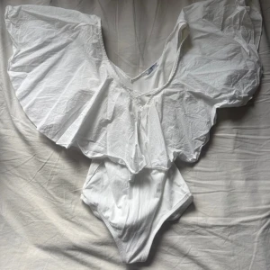 Vit volangbody från Zara  - Supersnygg vit body från Zara Trafaluc med stora volangärmar och v-ringning. Den har en omlottkänsla upptill och är tillverkad i mjuk bomull. Perfekt för dig som vill sticka ut med en unik och luftig topp i sommar.