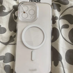 Transparent MagSafe-skal Holdit iPhone 15 Pro Max - Säljer ett transparent MagSafe-kompatibelt mobilskal från Holdit till iPhone 15 Pro Max. Skalets design är stilren med en vit cirkel på baksidan för MagSafe-funktion. Tillverkat i tålig plast som skyddar mobilen utan att dölja dess färg.