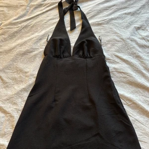 Svart halterneck kortklänning - Snygg svart kortklänning med halterneck och djup v-ringning. Klänningen har knyt i nacken och är ärmlös, vilket ger en stilren och trendig look. Perfekt för dig som vill sticka ut med en enkel men elegant stil.