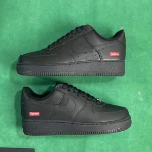 Nike Air Force 1 x Supreme Svart - Nike Air Force 1 Low i samarbete med Supreme. Helt svarta sneakers i skinn med klassisk siluett, Supreme-logga i rött på sidan och perforerad tå. Platt sula och snörning framtill. En ikonisk modell för dig som gillar streetwear.