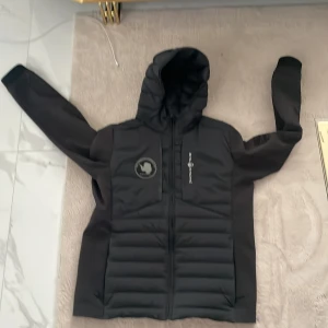 Svart Sail Racing pufferjacka 170 - Svart pufferjacka från Sail Racing med huva och coola Sail Antarctica Expedition-patchar på bröstet och ärmen. Jackan har quiltade partier framtill, hel dragkedja och lång ärm. Perfekt för dig som gillar stilren och sportig look.