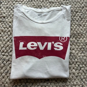 Vit Levi's t-shirt med röd logga - Vit t-shirt från Levi's med klassisk röd logga framtill. T-shirten har rund halsringning och korta ärmar. Tillverkad i mjuk bomull och har en liten röd Levi's-tag i sidan. Perfekt för en clean och avslappnad stil.