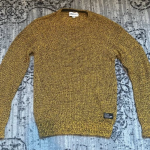 Gulmelerad stickad tröja D.M.D - Stickad tröja från D.M.D by denim day i gulmelerad färg med svarta inslag. Klassisk rund halsringning och långa ärmar. Perfekt för dig som gillar en chill och snygg stil. Liten svart patch nertill som detalj.