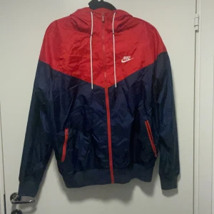 Nike vindjacka röd och marinblå - Nike vindjacka i rött och marinblått med huva och vita snören. Jackan har dragkedja framtill, två fickor med röda dragkedjor och elastiska muddar. Perfekt för dig som gillar sportig stil och vill sticka ut med färg. Helt ny använt Max 2 gånger.
