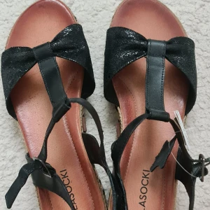 Svarta sandaler med rosett och flätad sula - Snygga svarta sandaler från Lasocki i äkta skinn med glittrig rosett framtill. Sulan är flätad i beige och svart för en cool espadrille-look. Remmarna är justerbara med spänne och sandalerna har platt sula. Perfekta för varma dagar och sommarens outfits.