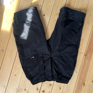 Svarta cargopants från Nike - Svarta cargopants från Nike med elastisk midja och benslut. Byxorna har stora fickor på sidorna och en liten vit Nike-logga på ena benet. Tillverkade i polyester, perfekta för en sportig och avslappnad stil.