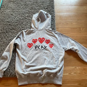 Grå hoodie Comme des Garçons Play - Grå hoodie från Comme des Garçons Play med dragkedja och huva. På bröstet finns det ikoniska röda hjärtat med ögon, och på ryggen flera röda hjärtan samt Play-loggan. Mjuk bomullskvalitet och känguruficka framtill.