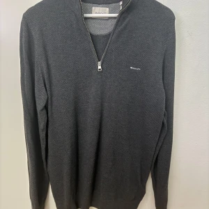 Mörkgrå half zip tröja från GANT - Snygg mörkgrå långärmad tröja från GANT med half zip och liten broderad logga på bröstet. Tröjan har en klassisk passform och är tillverkad i ett mjukt stickat material som känns skönt mot huden. Perfekt för lager-på-lager och stilren look.