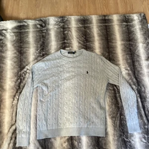Ljusgrå kabelstickad tröja Polo Ralph Lauren - Säljer en ljusgrå kabelstickad tröja från Polo Ralph Lauren. Tröjan har rund hals, lång ärm och klassisk broderad logga på bröstet. Ribbstickade muddar vid ärmslut och nederkant. Perfekt för dig som gillar stilren och tidlös look.