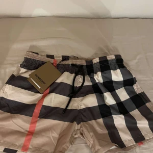 Burberry rutiga shorts med snörning - Snygga shorts från Burberry med klassiskt rutigt mönster i beige, svart, vitt och rött. De har elastisk midja med svart snörning och är perfekta för sommaren. Ikonisk design som sticker ut och passar dig som gillar exklusiva märken.