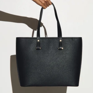 Svart shoppingväska i skinn - Stilren svart shoppingväska med två långa handtag och diskreta guldfärgade detaljer. Väskan är rymlig och har en klassisk, fyrkantig form som passar perfekt för att bära med sig allt man behöver. Tillverkad i skinn med en lätt glansig finish.