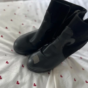 Svarta boots med klack och swirl-detalj - Svarta boots med hög klack och swirl-formad detalj på sidorna. Skorna har dragkedja på insidan och är gjorda i en mix av skinn och mocka. Rund tå och platt sula framtill. Perfekta för dig som gillar en edgy look.