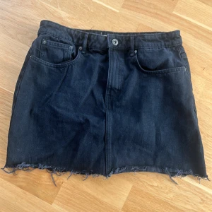 Svart jeanskjol med fransig kant  - Cool svart jeanskjol med rå, fransig nederkant och klassiska fickor fram. Kjolen har bälteshällor och knappstängning. Perfekt för dig som gillar en avslappnad och edgy stil.