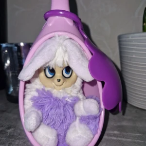 Kanin - Bilderna visar en söt, fluffig kaninliknande leksak med lila och vit päls, stora öron och blå ögon. Den kommer med ett äggformat, lila och rosa fodral som kan öppnas och stängas. Perfekt för dig som gillar gulliga samlarfigurer eller vill ha en unik dekoration.