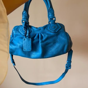 Blå skinnväska Marc by Marc Jacobs - Vintage blå handväska från Marc by Marc Jacobs i äkta skinn. Väskan har både handtag och justerbar axelrem, snygga detaljer och en metallbricka framtill. Fodret är mönstrat med stora bokstäver och väskan stängs med dragkedja.