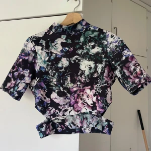 Blommig croppad topp med hög krage - Snygg croppad topp med blommigt mönster i lila, grön, vit och svart. Toppen har korta ärmar, hög krage och coola symmetriska detaljer vid midjan. Perfekt för dig som vill sticka ut med en färgstark och unik stil.