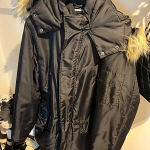 Svart parkajacka med fuskpäls - Svart parkajacka från Modernis med stor huva och fuskpälsdetaljer. Jackan har flera fickor, dragkedja och tryckknappar framtill. Perfekt för kalla dagar med sin långa passform och varma känsla.