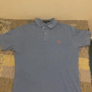 Blå pikétröja från Ralph Lauren - Snygg blå pikétröja från Ralph Lauren med klassisk krage och två knappar framtill. Tröjan har korta ärmar och den ikoniska orangea polospelaren broderad på bröstet. Tillverkad i mjuk bomull som känns skön mot huden. Skicket är 9/10 den är väldigt fräsch nu på sommaren, passar nästan till allt.