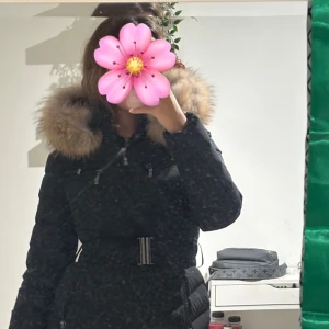 Svart dunjacka med pälsluva och bälte - Säljer en svart dunjacka från Hollies med stor fluffig päls runt luvan och markerad midja med bälte. Jackan har dragkedjor framtill och flera fickor, perfekt för kalla dagar. Snygg och stilren modell som håller dig varm hela vintern.