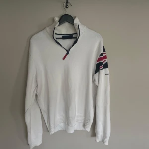 Vit stickad half zip-tröja Henri Lloyd - Vit stickad tröja från Henri Lloyd med half zip och hög krage. Union Jack-flagga på ena ärmen och broderad logga. Långärmad modell med ribbade muddar och nederkant. Perfekt för dig som gillar klassisk och sportig stil.
