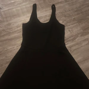 Svart ärmlös kortklänning - En stilren svart kortklänning med breda axelband och rundad halsringning. Klänningen har en figurnära överdel och lätt utställd kjol, vilket ger en klassisk siluett. Perfekt för dig som gillar enkel och tidlös design.