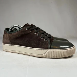 Grisch skor - Snygga bruna sneakers i mocka med mörkgrå lackad tå och matchande snörning. Skorna har en vit platt sula och rund tå, vilket ger en clean och modern look. Perfekta för dig som gillar stilrena detaljer och kontrasterande material.