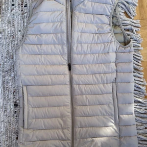 Beige quiltad dunväst med dragkedja - Snygg beige dunväst med quiltad design och hel dragkedja framtill. Västen har två fickor på sidorna och en stilren, enkel look. Perfekt att slänga över en hoodie eller tröja för extra värme. Passar både till jeans och mjukisbyxor.