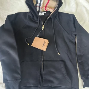 Svart Burberry hoodie med dragkedja - Helt ny svart hoodie från Burberry i bomull med klassiskt rutigt foder i huvan. Tröjan har dragkedja framtill, snörning i huvan och två stora fickor. Guldiga detaljer på dragkedjan och snörändarna ger en lyxig känsla. Perfekt för dig som gillar stilrena streetwear-plagg. Storlek M, men passar någon som har storlek S. 