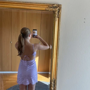 Ljuslila kort klänning med broderi - Superfin ljuslila kort klänning med tunna axelband, öppen rygg med snörning och v-ringning framtill. Klänningen har broderade blommor och håldetaljer längs nederkanten och bysten. Perfekt för sommarens varma dagar!