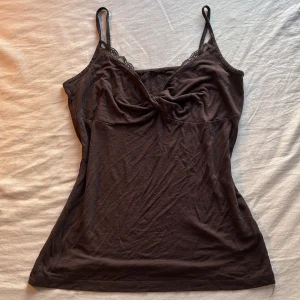 Brun linnetopp med spetsdetaljer - Snygg brun linnetopp med tunna axelband och spetsdetaljer vid urringningen. Toppen har en twistad detalj framtill som ger en cool look. 