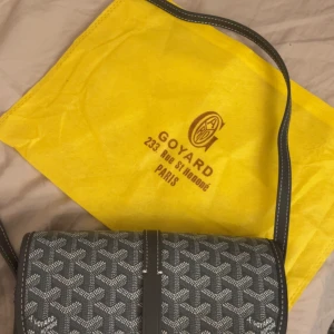 axelväska med monogram - Säljer en snygg axelväska  i klassiskt svart och grått monogrammönster. Väskan har justerbar axelrem, silverfärgad spänndetalj och insida i gul textil. Tillverkad i Frankrike med detaljer i skinn och ikoniskt mönster.
