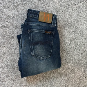 Mörkblå jeans från Nudie Jeans  - Snygga mörkblå jeans från Nudie Jeans Co med klassisk femficksdesign och slitningar för en cool look. Jeansen har orangea detaljer och patch bak i midjan. Perfekta för dig som gillar en tidlös och avslappnad stil.
