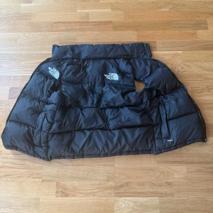Svart dunväst från The North Face XS - Säljer en svart dunväst från The North Face i storlek XS. Västen är quiltad med tydliga paneler, har dragkedja och innerficka med zip. Klassisk logga på bröstet och axeln. Perfekt för lager-på-lager och riktigt varm med 700 fill power.