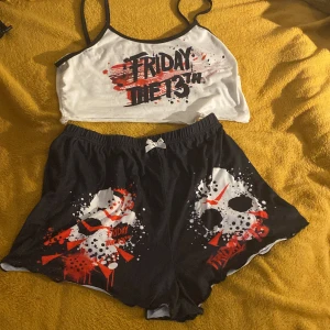 Friday the 13th shorts med tryck - Svarta shorts med elastisk midja och vit rosett framtill. Tryck med Friday the 13th och Jason-mask i vitt, rött och svart, samt coola färgstänk. Perfekta för dig som älskar skräckfilmer och vill ha en chill och edgy stil.