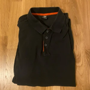 Svart pikétröja Helly Hansen  - Svart pikétröja från Helly Hansen Workwear i storlek XL. Klassisk krage, tre knappar framtill och orange detalj på insidan av kragen. Tillverkad i bomull, kortärmad och med diskret HH-logga nertill. Perfekt för en clean och enkel look.