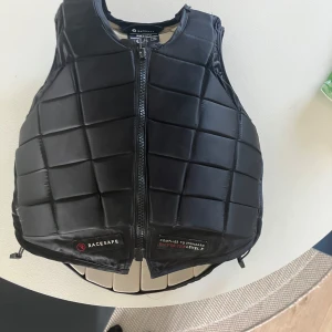 Svart skyddsväst - Skyddsväst från Racesafe, modell RS2010, i svart med quiltad design och dragkedja framtill. Västen har justerbara sidor med snörning. Den är använd en del men inget som syns och har inga större skador, storlek L i barn ca xxs 