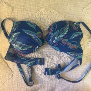 Blå bikiniöverdel med tropiskt mönster - Snygg blå bikiniöverdel med tropiskt bladmönster i grönt, turkost och orange. Justerbara axelband och klassisk knäppning bak. Perfekt för strandhäng och poolparty. Tillverkad i mjukt syntetmaterial som torkar snabbt.