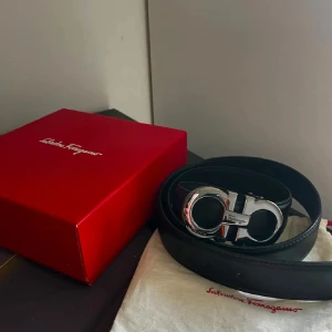 Svart skärp från Salvatore Ferragamo - Snyggt svart bälte från Salvatore Ferragamo med ikoniskt silverfärgat spänne. Bältet är gjort i slätt skinn och har en stilren, klassisk design som passar till många outfits. Kommer med originalförpackning och dustbag.