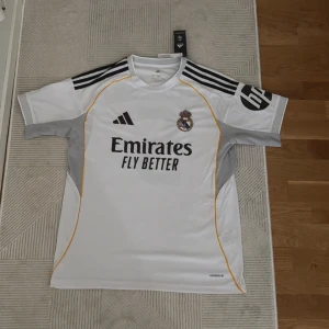 Real Madrid Mbappé 9 Adidas tröja - Snygg vit Real Madrid fotbollströja  med Mbappé och nummer 9 på ryggen. Tröjan har svarta detaljer, gula linjer och grå partier på sidorna. Klassisk kortärmad modell med klubbmärke och sponsortryck framtill. Tillverkad i lätt och ventilerande material.