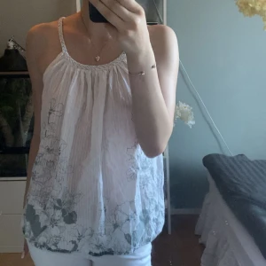 Vit blommig linnetopp från Monode - Supersöt vit linnetopp från Monode med tunna flätade axelband och blommigt mönster i grått. Toppen är luftig och har en lätt rynkad effekt upptill. Perfekt för varma dagar och ger en fräsch och somrig vibe.