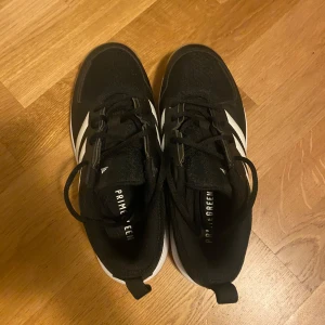 Svarta Adidas sneakers med vita ränder - Snygga svarta Adidas sneakers med klassiska vita ränder på sidorna och gummisula i ljusbrun färg. Ovandelen är i textil och syntet, med snörning och låg profil. Perfekta för dig som gillar sportig stil och vill ha bekväma skor till vardagen.